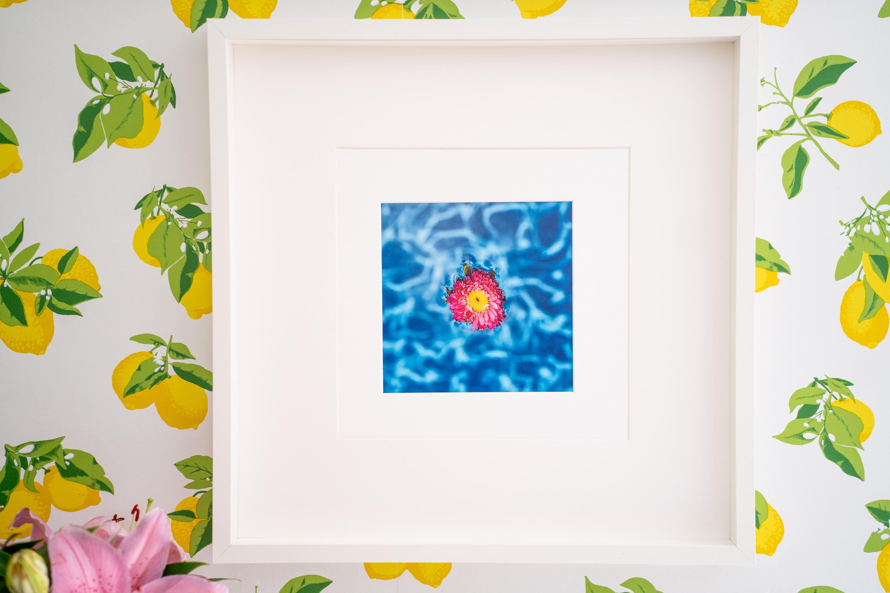A framed fine art print featuring a vibrant pink and yellow chrysanthemum floating on a shimmering blue pool, displayed on a wall with playful lemon-patterned wallpaper. The bright and refreshing composition brings a summery, tropical feel to the space.