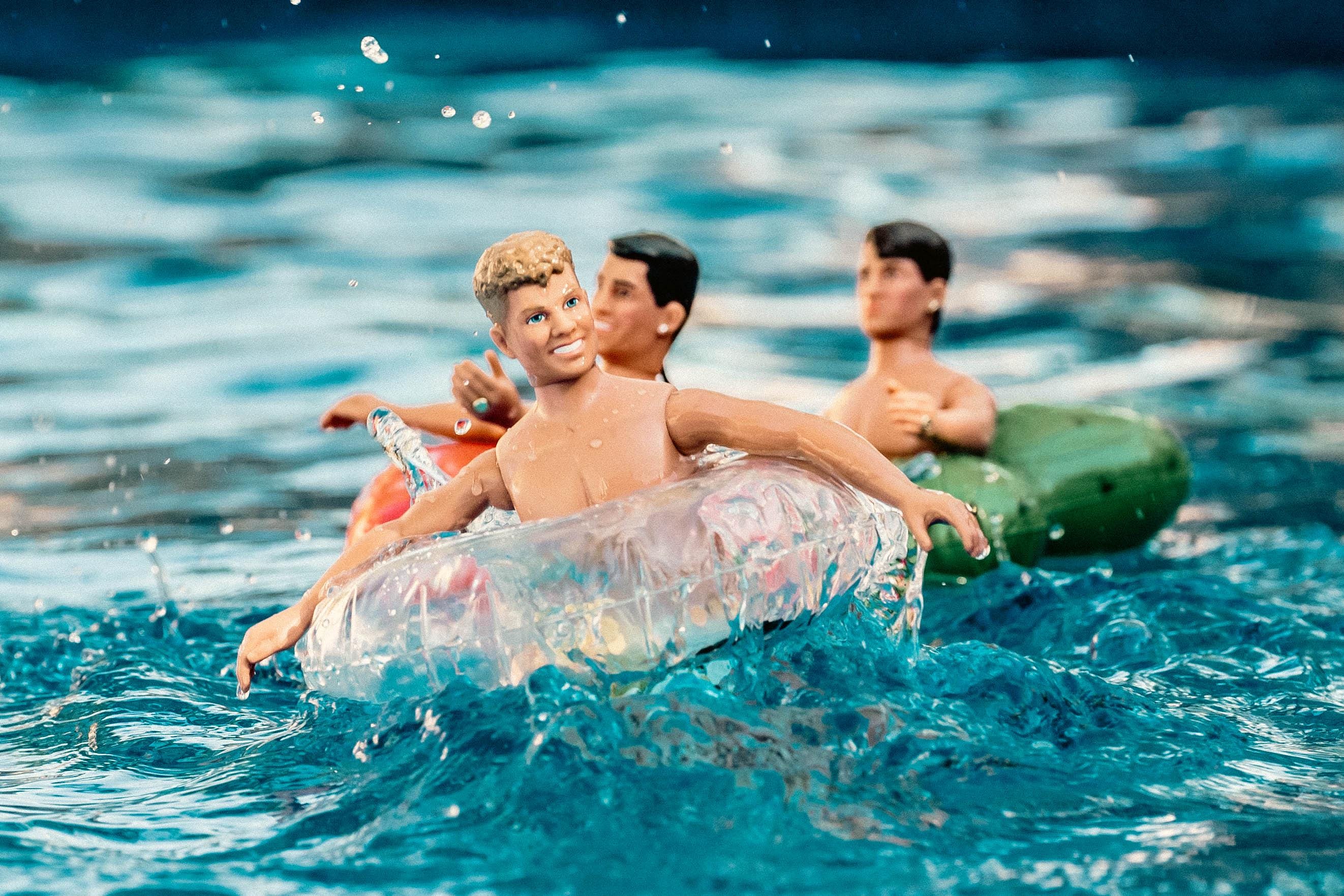 Three vintage New Kids on the Block dolls floating on inflatable pool rings in splashing blue water, with bright summer lighting and playful retro vibes
