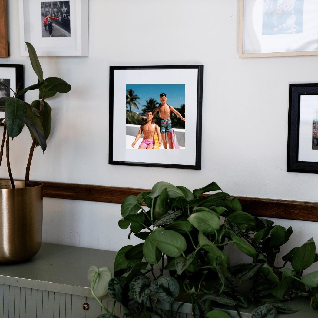 A framed fine art print featuring vintage New Kids on the Block dolls, Danny and Jordan, styled in retro swimwear and posed in a tropical rooftop scene with palm trees in the background. Danny is lounging on a yellow beach chair wearing pink shorts, while Jordan stands confidently, sporting sunglasses, a braid, and patterned swim trunks. The framed print is displayed in a modern interior setting, surrounded by lush green plants