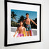 A framed fine art print featuring New Kids on the Block dolls, Danny and Jordan, in a playful, sun-soaked rooftop scene in a tropical setting. Danny, seated in a yellow lounge chair, wears pink shorts with his signature long hair, while Jordan stands beside him sporting sunglasses, a side braid, and patterned swim trunks. Palm trees sway in the background, adding to the retro summer vibe.