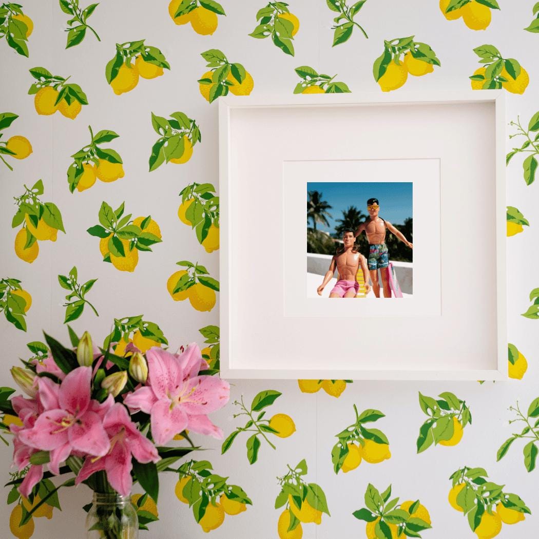 A framed fine art print featuring vintage New Kids on the Block dolls, Danny and Jordan, posed in a tropical rooftop scene with palm trees and a bright blue sky. Danny relaxes on a yellow beach chair wearing pink shorts, while Jordan stands confidently in sunglasses, a braid, and patterned swim trunks. The artwork is displayed on vibrant lemon-patterned wallpaper, with fresh pink lilies adding a lively and nostalgic touch, making this a perfect statement piece for NKOTB fans and collectors.