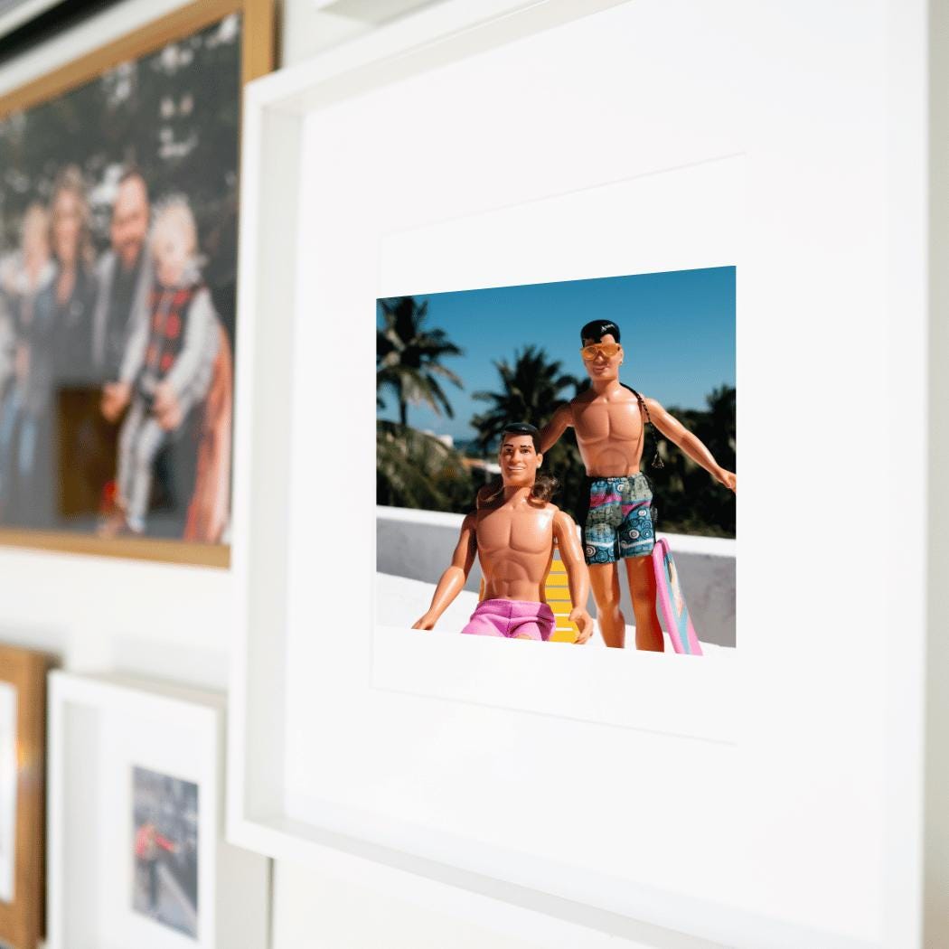 A framed fine art print featuring vintage New Kids on the Block dolls dressed in retro swimwear, posing in a tropical rooftop setting with palm trees and a bright blue sky in the background. One doll lounges on a yellow beach chair while the other stands confidently, wearing sunglasses and a stylish braid. The playful nostalgia and vibrant colors make this a unique and bold statement piece for pop culture and retro collectors.