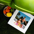 The image shows a printed photograph of two male dolls posed in a sunny, tropical setting. The dolls are dressed in beachwear, with one wearing pink swim trunks and the other in patterned shorts, sunglasses, and a crossbody bag. The printed photo is placed on artificial grass, with a bowl of citrus fruits (oranges, lemons, and limes) nearby. A portion of a green inflatable or chair is visible in the top part of the frame, adding a playful and summery vibe to the composition.