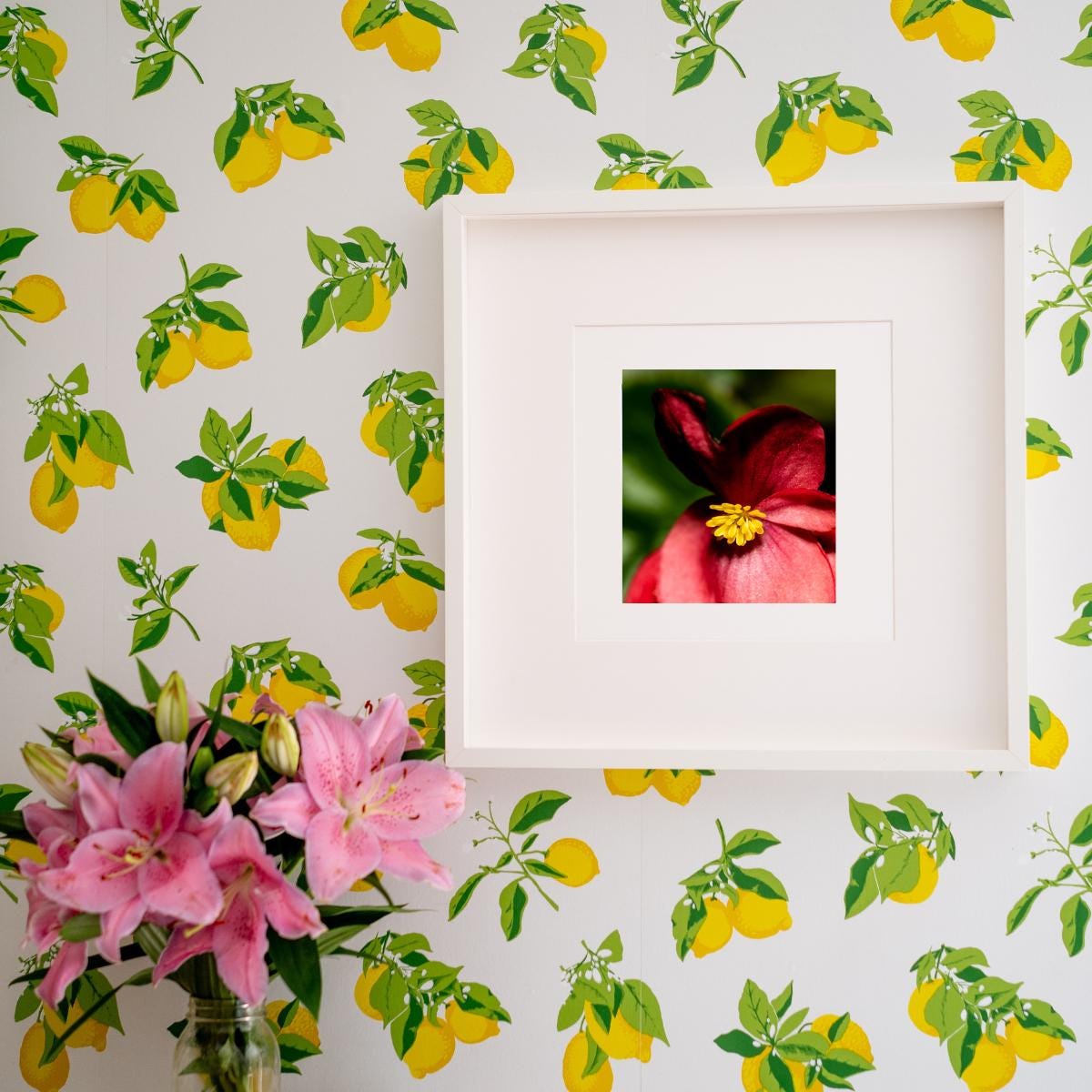 A framed fine art print featuring a macro photograph of a red begonia with a bright yellow center, set against a lush green background. The artwork is displayed on a vibrant lemon-patterned wallpaper, complemented by a vase of fresh pink lilies. The combination of bold floral elements and cheerful decor creates a lively and elegant interior setting.