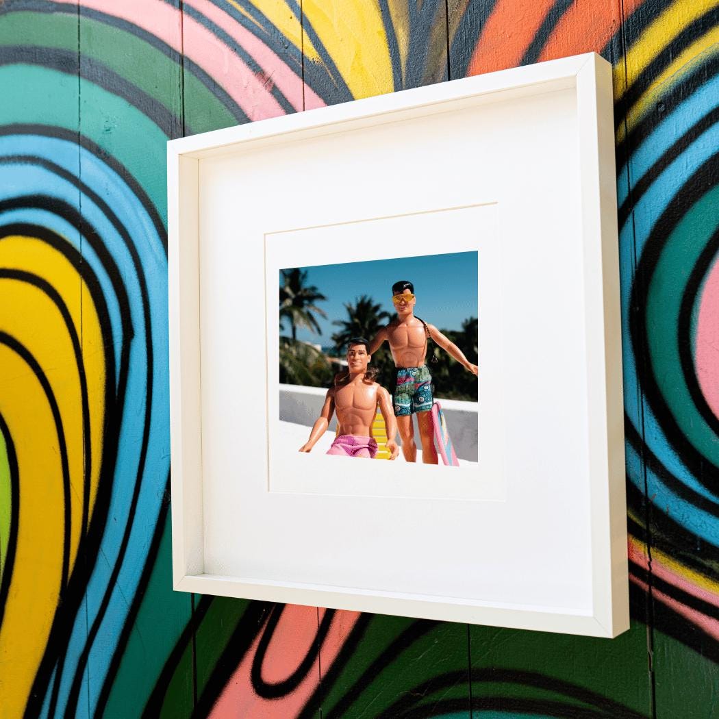 A framed fine art print featuring vintage New Kids on the Block dolls, Danny and Jordan, dressed in retro swimwear and posing in a tropical rooftop setting. Danny lounges on a yellow beach chair in pink shorts, while Jordan stands confidently wearing sunglasses, a braid, and patterned swim trunks. The vibrant graffiti wall in the background adds a bold, artistic touch, making this a playful and nostalgic statement piece for NKOTB fans and collectors.