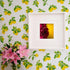 A framed fine art print featuring a striking macro photograph of a deep magenta dahlia set against a golden-yellow background. Displayed on a lemon-patterned wallpaper, the artwork adds a bold contrast to the fresh and lively decor. A bouquet of pink lilies in a glass vase enhances the vibrant and nature-inspired aesthetic of the space.