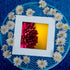 A framed fine art print of a deep magenta dahlia set against a golden-yellow background, displayed on a vibrant blue metal table. Surrounding the artwork is a delicate arrangement of pale white chrysanthemums, creating a visually striking contrast between the warm floral tones and the cool blue setting. A serene and artistic composition perfect for floral and nature-inspired decor.