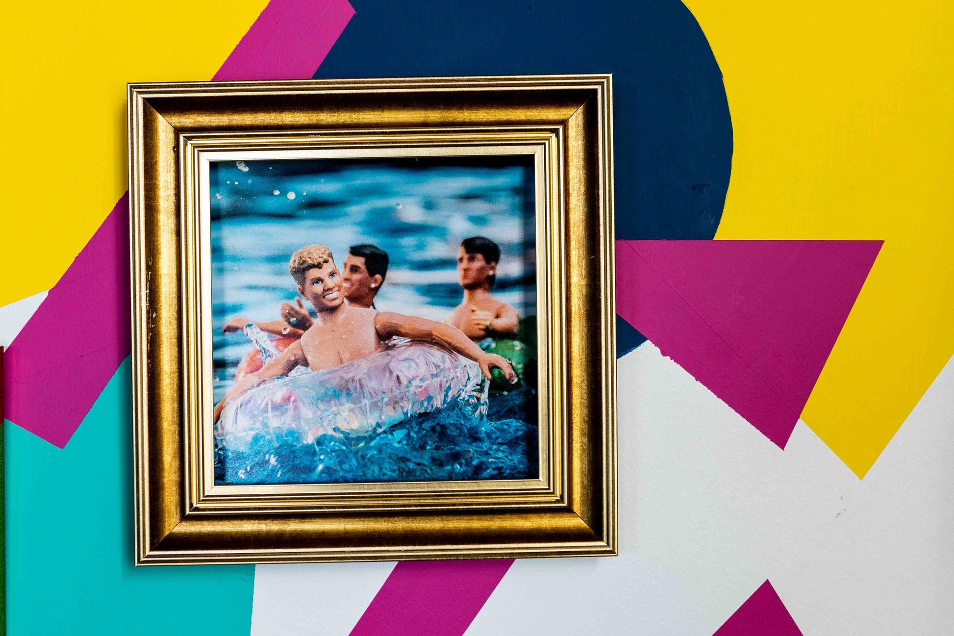 Three vintage New Kids on the Block dolls floating on inflatable pool rings in splashing blue water, with bright summer lighting and playful retro vibes in gold frame on bright mural wall.