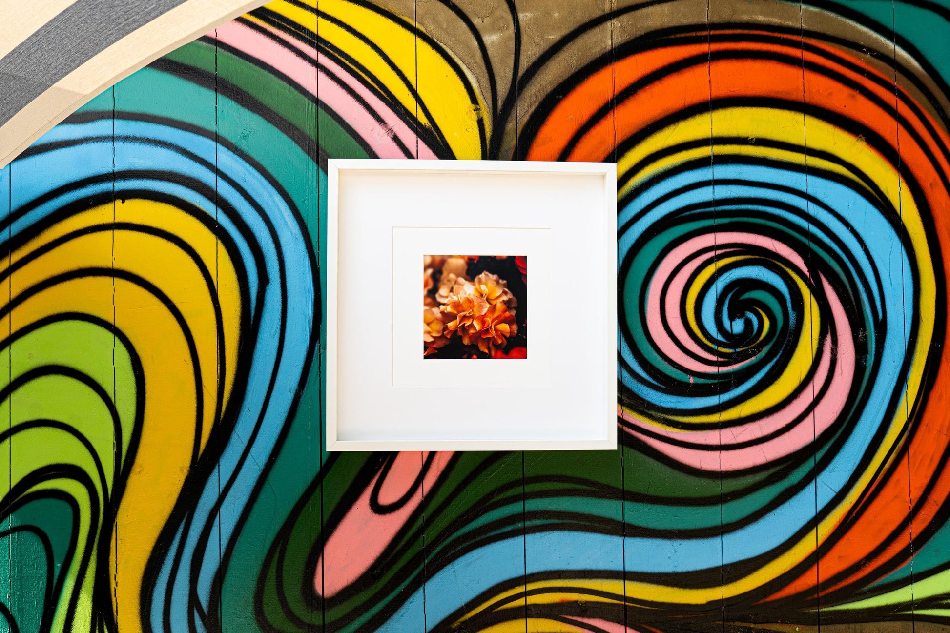A framed floral art print featuring golden-orange flowers is displayed against a vibrant, multicolored swirl mural. The striking contrast between the warm floral tones and the bold, abstract background enhances the artistic appeal.